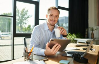 Handsome young man with a cup of coffee sits at his modern office desk using a tablet while smiling.